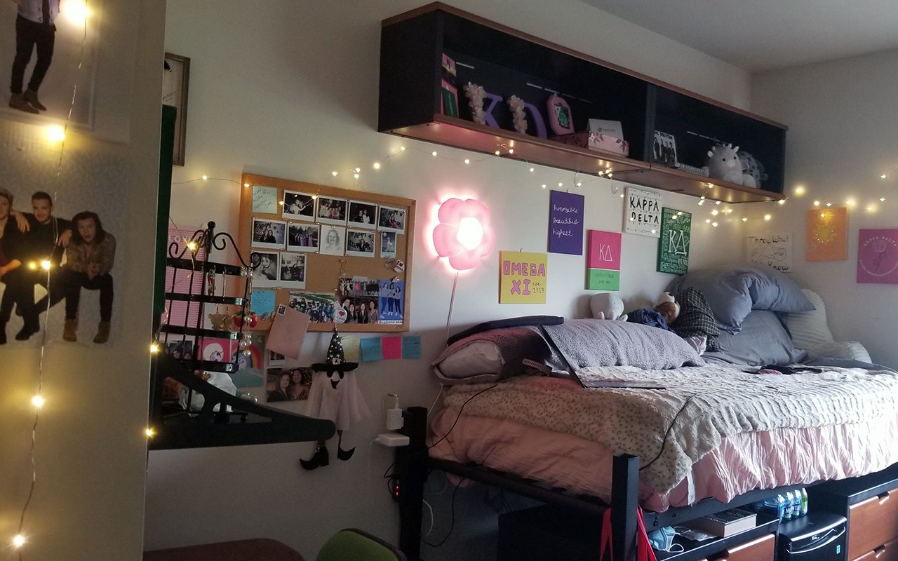 residence hall bedroom featuing many pictures and posters on the wall and the shelf above the bed. strands of lights are hung above bed  