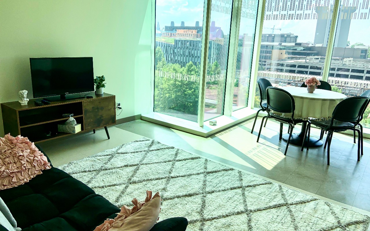 A table with chairs, a television, and a couch are located in a large room with large windows. 