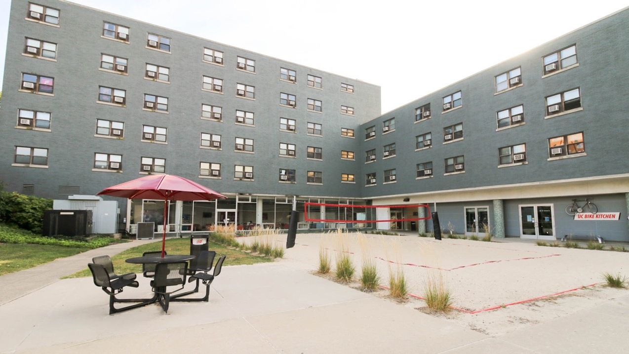 Exterior of Dabney Hall. There is a sand volleyball court and a lounge area that has a table with and umbrella and four chairs.